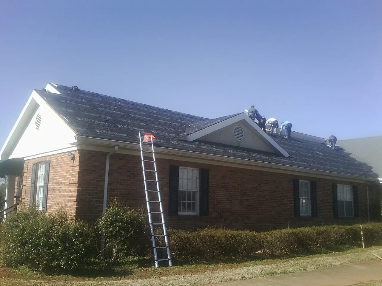 Crew tearing shingles off a brick ranch roof with a ladder set — Metal Roof Repair work by our crew in Muldrow