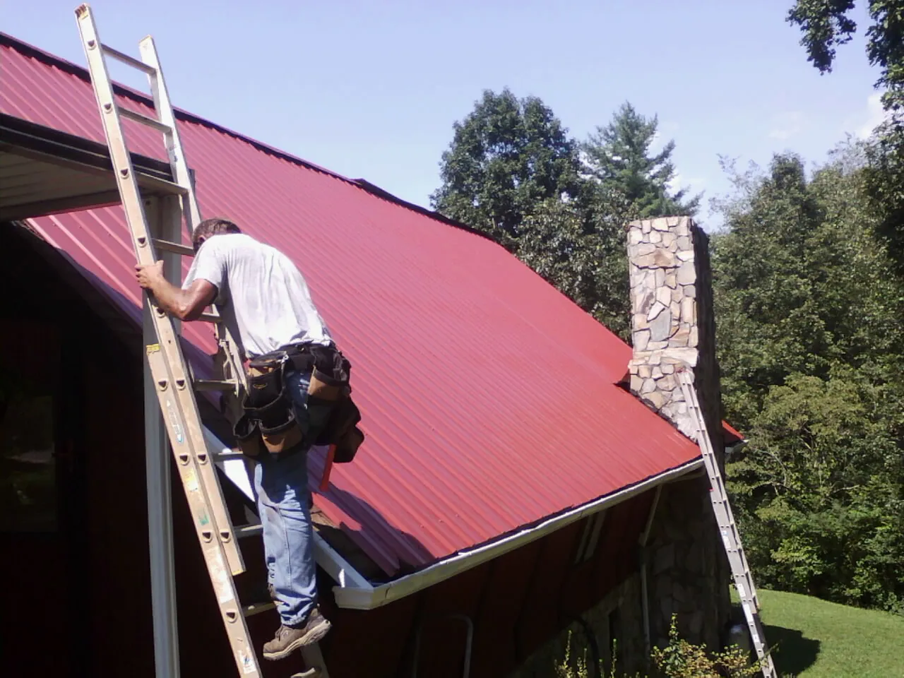 Roofer on a ladder beside a red metal roof and stone chimney — Soffit & Fascia Repair by Roofing Brothers in Muldrow